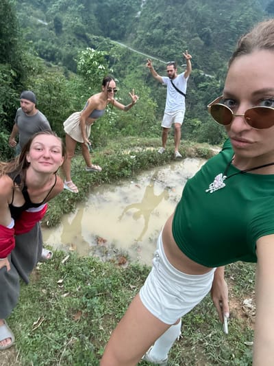 A group of five individuals is captured in an outdoor selfie-style photograph, taken in what appears to be a lush, mountainous region of Nậm Dịch Commune, Vietnam. The image is oriented with the selfie-taker at the top, looking down into the camera, capturing the group and the surrounding environment.
The main subject, a woman with blonde hair wearing sunglasses, a green crop top, and white shorts, holds the camera for the selfie. She has a distinct butterfly-shaped necklace. Below her, four other individuals are visible. To the right, a man in a white t-shirt and white shorts is making peace signs. To his left, another woman in a grey top and light-colored skirt is also posing with peace signs. In the bottom left, a woman in a red top and black tank top smiles towards the camera, alongside a man in a grey t-shirt and cap.
The setting is characterized by dense green foliage and trees, indicating a natural or rural landscape. A prominent feature in the midground is a large, muddy puddle, reflecting the shapes of some of the individuals, particularly the man in white and the woman in grey. The background shows distant, hazy green mountains or hills under what appears to be an overcast sky, suggesting diffused daylight.
The group appears engaged in a casual outdoor activity, likely hiking or exploring, and is pausing to take a souvenir photo. Their expressions are generally positive and relaxed, with smiles and playful poses. There is no visible text in the image.