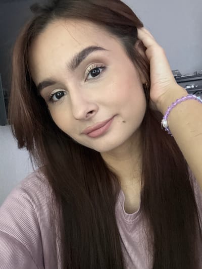 A close-up, medium-shot photograph shows a young woman with long, dark brown hair, who is looking directly at the camera. Her hair is parted on the left and frames her face. She has light brown eyebrows, dark eyes with visible eyeliner and mascara, and a neutral expression. Her lips are lightly colored with lipstick. A faint mole is visible on her right cheek. She is wearing a light pink, ribbed t-shirt. A purple beaded bracelet adorns her left wrist. Her right hand is raised, with her fingers gently touching her hair near her temple. The background is a blurred, neutral gray, suggesting an indoor setting, possibly a room. There are no visible elements that place the image in the city of Kuyuki, Russia.
