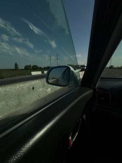 This media file is a photograph taken from inside a car, looking out the driver's side window and rearview mirror. The perspective is rotated, suggesting the car is either parked at an angle or the photo was taken with the camera tilted.
The scene outside the car appears to be a rural or semi-rural area. Through the window, a cloudy sky with patches of blue is visible, along with some power lines stretching across the frame. Below the sky, there's a glimpse of trees and what looks like agricultural fields. In the distance, a white truck is visible, possibly on a road. The rearview mirror reflects a road and some of the surrounding landscape, including more power lines.
The interior of the car is also visible, with the black textured door panel, a silver door handle, and part of the dashboard. A portion of the car's frame or pillar is also present in the foreground. The lighting suggests it could be daytime, possibly late afternoon or early morning given the quality of light and the contrast between bright sky and darker interior.
No people are visible in the image. The overall impression is a snapshot of a travel moment, capturing the view from a moving vehicle. The location context of Istiqlol, Uzbekistan, indicates the geographical setting of this scene. There is no visible text in the image.