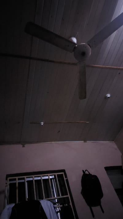 The image depicts an interior view of a room in New Nyanya, Nigeria, characterized by a wooden slatted ceiling and a pinkish-purple wall. A ceiling fan with three blades is prominently featured, its central housing and motor visible. One blade is angled downwards, obscuring a portion of the ceiling. Two light fixtures are mounted on the ceiling, one to the left and another to the right, both appearing to be bare bulb holders. A dark, rectangular object with vertical bars, likely a window grate, is positioned on the lower portion of the wall, with some white and dark fabric draped in front of it. A black backpack is hanging on the wall to the right of the window. The lighting in the room is dim, creating shadows and highlighting the textures of the ceiling and walls.
