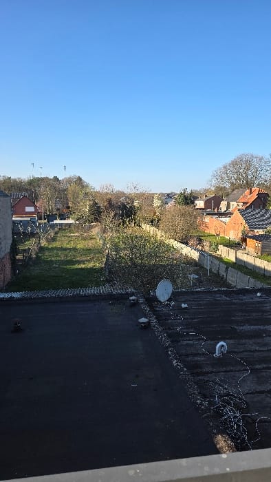 An elevated view from a building in Heusden, Belgium, captures a residential area under a clear, cloudless blue sky. The immediate foreground features a light grey concrete ledge at the bottom edge, overlooking a dark, flat-membrane roof. On the roof, a white parabolic satellite dish is positioned towards the center-right. To the left, two dark-colored, circular ventilation structures are visible. To the right, a coiled mass of dark cables and a curved white pipe lie on the roof surface.

The midground is dominated by an irregular, rectangular grassy plot of land, appearing unmaintained with visible weeds and scattered small deciduous trees and shrubs, some showing early foliage. This plot is bordered on the left by a chain-link fence and on the right by a concrete panel fence constructed of vertical posts and horizontal slabs.

In the background, a residential neighborhood extends, characterized by various houses with red-tiled roofs and brick facades. On the far left, a distinct red-brick house with a white roller shutter covering a window is visible. Numerous deciduous trees, some still largely bare, are interspersed among the houses and form distant tree lines. Above the trees on the far left horizon, two tall, dark, pole-like structures are present. No individuals are visible in the scene. The entire environment is illuminated by natural daylight.