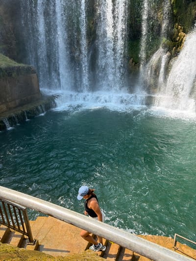 A woman wearing a black tank top, white shorts, a blue baseball cap, and sneakers descends a stone staircase next to a large waterfall in Jajce, Bosnia and Herzegovina. The waterfall cascades over a rocky cliff face covered in greenery into a pool of clear, blue-green water. The staircase has a metal railing and is illuminated by sunlight. The overall scene is natural and serene.