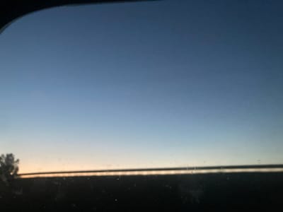 The image captures a twilight sky over a dark, silhouetted landscape. The sky transitions from a deep blue at the top to a lighter blue and then a soft orange-yellow hue near the horizon, suggesting either sunrise or sunset. A thin, dark horizontal line separates the sky from the foreground, likely representing the horizon or a distant feature. On the left side of the image, a blurry, dark shape resembling a tree or a clump of foliage is visible against the lighter sky. The overall impression is one of a serene, natural environment. The image does not provide specific visual cues to pinpoint its exact location within Ракітніцкі сельскі Савет, Belarus, beyond it being an outdoor scene likely in a rural or semi-rural setting.