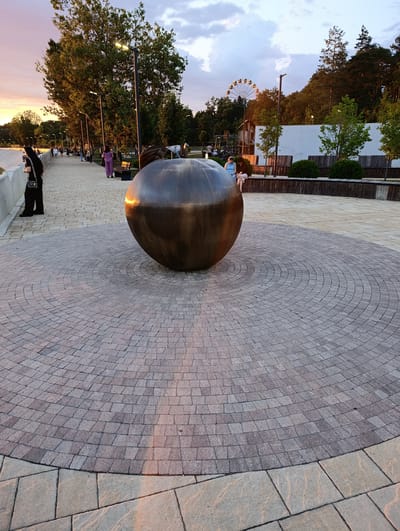 This image depicts an outdoor scene at dusk in a public park or promenade, potentially in the city of Kelermesskaya, Russia. The central focus is a large, spherical, metallic sculpture situated on a circular paving stone pattern. The sculpture reflects the warm hues of the setting sun. To the left, a paved walkway is lined with trees and lampposts. Several individuals are walking or sitting along the path, some engaged in conversation. In the background, a Ferris wheel is visible, suggesting the presence of an amusement area or a larger city park. The overall atmosphere is serene and suggests a leisurely evening.