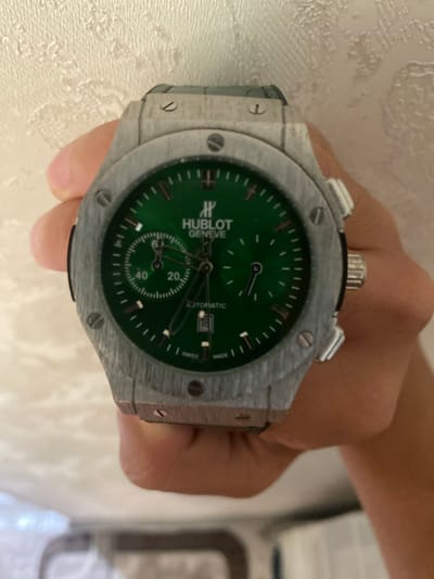 A person's hand holds a Hublot watch with a green dial. The watch has a silver-toned metal case and a green inner bezel. The dial features silver-toned hour markers and hands, with a date window displaying "18" at the 6 o'clock position. A subdial with a smaller hand is visible at the 9 o'clock position. The watch is being held in front of a textured, off-white background. The image is cropped closely to the watch and hand, obscuring the person's face and body. There is no discernible information to indicate a location in Istaravshan, Tajikistan.