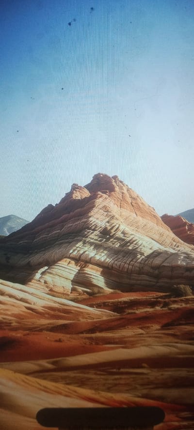 The image depicts a desert landscape with layered sandstone formations bathed in sunlight. The formations exhibit a gradient of colors from reddish-brown in the foreground to lighter tan and white further back, with distinct horizontal striations indicating geological layers. The sky above is clear and a pale blue, transitioning to a lighter hue near the horizon. The foreground shows rolling desert terrain with patches of vegetation. There are no people or man-made objects visible in the natural landscape. The image does not provide any specific information or visual cues that would indicate a location in Kudymkar, Russia, as the landscape is characteristic of arid desert regions.