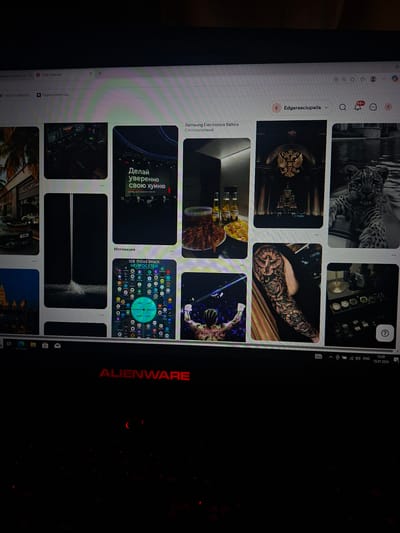 The image shows a close-up of a computer monitor displaying a Pinterest board. The Pinterest board features a grid of images, including a man in a boxing ring, a full sleeve tattoo of a dove and roses, a collection of watches, a leopard, a waterfall, and a group of bottles and snacks. The monitor is an Alienware brand, indicated by the red logo. The bottom of the screen shows the Windows taskbar with the time and date "13:09 19.07.2026". The background is dark and out of focus. There is no visible indication of the city of Užuežerė, Lithuania in the image.