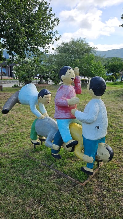 The image shows a playful outdoor sculpture of children in a park in Geoje-si, South Korea. The sculpture depicts at least four figures, appearing to be children, engaged in some form of game or interaction. One figure, wearing a pink shirt and blue pants, is laughing with its hand raised to its face. Another figure in a white shirt and blue pants stands opposite, facing the laughing figure. A third figure, in a light blue shirt and gray pants, is positioned lower and appears to be in motion, possibly climbing or being carried. A fourth figure, primarily yellow with a black head, is partially visible at the bottom, suggesting it is part of the base or a supporting element of the sculpture. The figures are stylized, with simplified features and bright, solid colors.
The sculpture is situated on a grassy area with scattered trees in the background. The ground is covered in green grass, with some patches of dry grass and fallen leaves, indicating an outdoor park setting. A few fallen branches are visible on the ground near the sculpture. In the distance, there are more trees, some low hills or mountains, and a building structure. The sky above is partly cloudy with patches of blue sky. The overall impression is that of a public art installation in a recreational area.