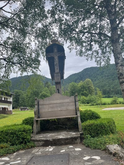 A religious monument featuring a crucifix and a wooden bench stands in a grassy area in Ettal, Germany. The monument is made of wood and stone, with the crucifix at the top, depicting Jesus. A wooden bench is positioned in front of the crucifix, with stone steps leading up to it. The scene is framed by trees and a cloudy sky. In the background, a building with white walls and brown shutters, and a forest-covered mountain can be seen. The overall impression is one of a serene, rural setting.