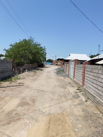 This is a vertical, eye-level, medium shot of a dirt road in a rural area. The road is unpaved and appears to be a residential street. On the left side of the road, there is a wall constructed of concrete blocks, followed by a wooden fence and a large green tree. On the right side of the road, there is a wall constructed of concrete blocks and red bricks, with a metal gate. In the background, there are several houses with white metal roofs. The sky is clear and blue. The scene suggests a typical day in a village, possibly in the city of Sovet, Kyrgyzstan, given the style of houses and fencing. There are no people visible in the image. The overall impression is one of a quiet, sun-drenched rural settlement.