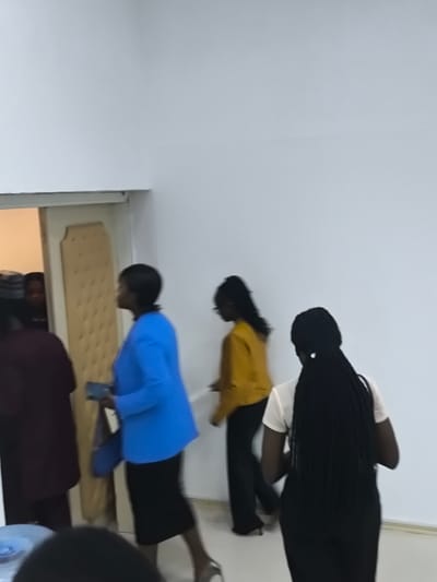 This image, set in Kızılbaş, Northern Cyprus/Cyprus, depicts an indoor scene with several individuals moving through a white-walled area. The environment consists of plain white walls and a partially open light-colored door with decorative paneling on the left, revealing a darker space beyond where a person's face is dimly visible.
Five individuals, all appearing to be of dark complexion, are in motion within the frame. From left to right:
1. A dark-skinned individual in a dark red/maroon top is partially visible from the back, positioned near or within the open doorway.
2. A dark-skinned woman walks across the mid-ground, wearing a bright blue blazer, a dark skirt, and light-colored high heels. She carries a dark blue clutch bag in her left hand. Her hair is short and dark.
3. Following her, a dark-skinned young woman in a mustard yellow long-sleeved top and dark trousers walks with glasses. Her long, dark hair is styled back.
4. In the right foreground, a dark-skinned young woman is captured from behind, facing away from the camera. She wears a light-colored short-sleeved top and dark trousers, and has long, dark braided hair.
5. A partial view of a head and shoulder belonging to another dark-skinned individual is visible in the bottom left corner, positioned near a light blue, possibly plastic, object.
The individuals are walking in various directions, primarily across the frame from left to right or towards the right side, suggesting movement through a passage or entry/exit from a room. There are no direct interactions captured. The image shows some motion blur, consistent with the subjects' movement.