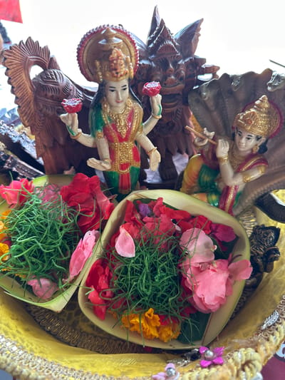 The image shows an offering arrangement, likely in Rendang, Indonesia, due to the Balinese cultural objects. The arrangement consists of two painted statuettes of deities, one playing a flute, mounted on a carved wooden base, and two small leaf-woven baskets filled with floral offerings, including rose petals and green grass-like plants. The entire arrangement sits on a yellow cloth with decorative trim.