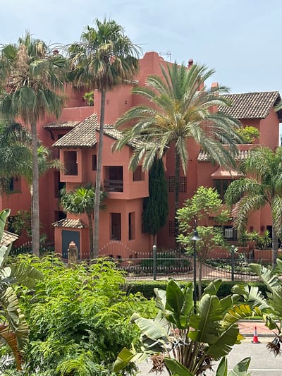 The image shows a close-up view of a reddish-orange building complex with several palm trees and lush green foliage in front of it. The building has tiled roofs and multiple balconies and windows. The environment appears to be a warm, sunny climate, consistent with a location in Resinera-Voladilla, Spain. No people are visible in the scene.