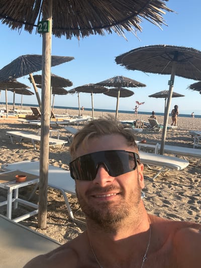 A man takes a selfie on a beach in Angelochori, Greece. He is wearing sunglasses and a silver chain necklace. Behind him, straw umbrellas shade rows of lounge chairs on the sandy beach. In the background, people are visible near the water, and a kite is flying in the sky. The scene is bathed in sunlight under a clear blue sky.