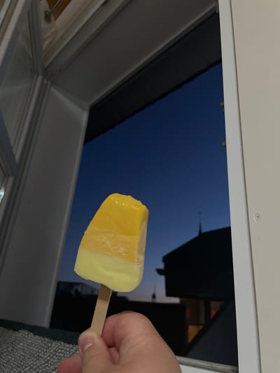 An individual's hand holds a yellow and white popsicle from the bottom, with the popsicle positioned in front of an open window. Through the window, a dark blue twilight sky is visible, with the silhouette of a building and a steeple in the background, possibly indicating a church. The scene is indoors, with the interior of a room or building visible in the foreground and to the left. The context suggests this may be a residential setting, with the window offering a view of the town of Waldbröl, Germany, at dusk.