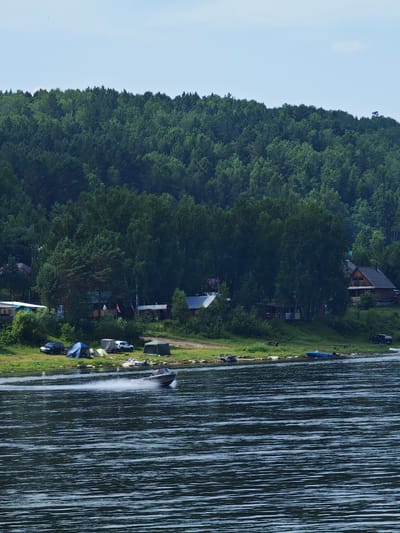 This image depicts a serene waterscape with a densely forested hillside in the background. In the foreground, a wide, dark body of water exhibits ripples and a subtle sheen, suggesting moderate movement. A small motorboat is actively navigating the water, leaving a white spray trail behind it. The shoreline, visible in the middle ground, features a grassy bank with several tents pitched, indicating a camping or recreational area. Various vehicles, including cars and what appears to be a recreational vehicle, are parked along the bank. A few rustic structures, likely cabins or houses, are nestled amongst the trees on the hillside. The overall environment is natural and suggests a peaceful, outdoor setting. The location is identified as Lugovskoe, Russia.