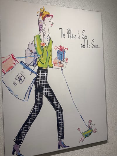 An illustrative artwork is mounted on a light-colored, textured wall in Bal Harbour Village, United States of America. The artwork depicts a stylized, slender woman in profile, walking towards the right side of the frame. She has blonde hair tied up, red-framed sunglasses pushed onto her head, and prominent red lipstick. Her attire includes a light green long-sleeved top, dark pants with a white grid pattern, and pointed-toe, bluish-purple high-heeled boots.
The woman is engaged in multiple actions: her right hand holds a purple, older-model cellular phone to her ear. Her left arm is laden with various items: a large white shopping bag featuring a dark blue logo (resembling a stylized 'B'), a smaller red-orange bag, and a light purple backpack slung over her shoulder. Her left hand also carries a stack of three gift boxes, with the topmost being a blue box adorned with white polka dots and a red bow, resting on two smaller rectangular boxes (one pink, one beige). A thin blue leash extends from her left hand downwards.
Following the leash, a small, stylized pink dog, wearing a green and yellow plaid coat, walks alongside the woman, also facing right. To the right of the woman, handwritten-style text reads: "The Place to See and be Seen...". The wall behind the artwork is a textured, off-white or light beige surface.