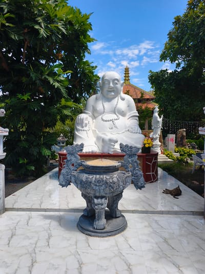 This is a full-shot, eye-level, outdoor photograph of a large white statue of the laughing Buddha, also known as Budai, situated in a temple complex in Tuy An Nam, Vietnam. The statue is the primary subject, depicted in a seated position with a serene, smiling expression and a beaded necklace. In front of the Buddha statue, on a marble floor, is a large, ornate incense burner with intricate dragon carvings, filled with sand and incense sticks.
To the right of the Buddha statue, a smaller white statue of a female figure stands, possibly a bodhisattva. Behind the main statue, a multi-tiered pagoda structure with an orange tiled roof is visible. The scene is framed by lush green trees and foliage, with a clear blue sky dotted with white clouds above. The ground is paved with white marble tiles.
On the far right side of the frame, a brown dog is lying down on the tiled floor. The overall atmosphere is peaceful and contemplative, typical of a religious site. The photograph is well-lit, suggesting it was taken during daylight hours.