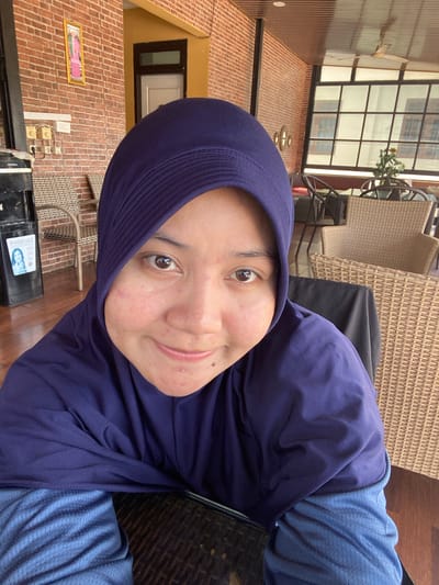A close-up selfie is taken by a person wearing a dark blue hijab and a blue shirt. The person's face is the primary focus, with their eyes looking directly at the camera and a slight smile. Their skin shows some blemishes and redness. In the background, there are elements of an outdoor seating area, likely a cafe or restaurant, with wicker chairs and tables visible. The architecture suggests an Indonesian setting, with brick walls and a covered ceiling. A water dispenser with an "Instaload" label is to the left of the frame, and a framed picture hangs on the brick wall above it. The overall lighting suggests it is daytime.
