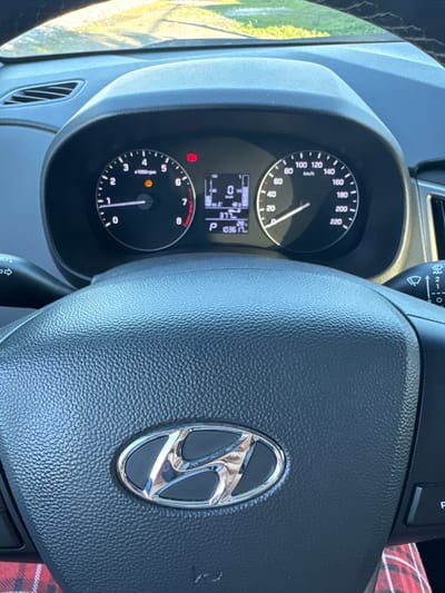The image shows the interior of a Hyundai car, focusing on the steering wheel and dashboard. The dashboard features a digital display with information such as speed (0 km/h), gear (P), and trip distance (10361 km). The tachometer shows 0 rpm, and there's an orange illuminated warning light, likely for the engine or tire pressure. The steering wheel has the Hyundai logo prominently displayed in the center. To the left of the steering wheel, a turn signal lever is visible, and to the right, a windshield wiper control stalk is present. The background outside the car is blurred but suggests a sunny outdoor environment with some greenery. The image is likely taken by someone in the driver's seat. The car is parked in Starokhalilovo, Russia.