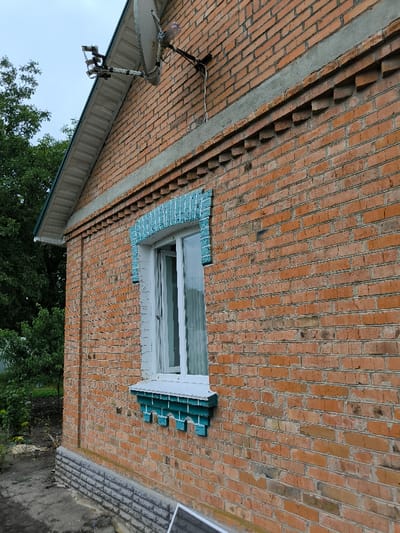 The image depicts the exterior side of a building, identified as being in Pysarivka, Ukraine. The structure is primarily constructed of red-orange brick, laid in a stretcher bond pattern, with a visible gable end featuring a white soffit and a dark green gutter. A light grey concrete band runs horizontally below the soffit, and beneath it, a decorative course of bricks is stepped out.

A white-framed window is centrally located on the brick wall, adorned with an arched surround and sill detail in a distressed teal-blue finish, featuring carved or molded patterns. The window's left pane is slightly ajar, and white interior blinds or curtains are visible through the glass. Below the window, the foundation of the building is faced with grey, textured blocks, suggesting a concrete or stone base.

High on the brick wall, above and to the right of the window, a light grey satellite dish with multiple LNBs is mounted. Wiring extends from the dish along the brickwork.

The ground immediately adjacent to the building is a patch of dark, disturbed earth, with some sparse green vegetation. In the lower foreground, a dark, rectangular object, possibly a solar panel or framed object, is partially visible on the ground. Mature green trees with dense foliage are visible in the background to the left, under an overcast sky. No people, animals, or active interactions are visible within the frame.