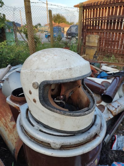 A weathered, white motorcycle helmet sits atop a rusty metal barrel in an outdoor area. The helmet is dirty and the interior lining is torn. The background shows a chain-link fence, wooden posts, overgrown vegetation, and a rustic wooden structure. In the distance, buildings, a blue truck, and a dark vehicle are visible. The scene appears to be in Babuk, Bulgaria, and suggests a junkyard or an abandoned outdoor storage area.