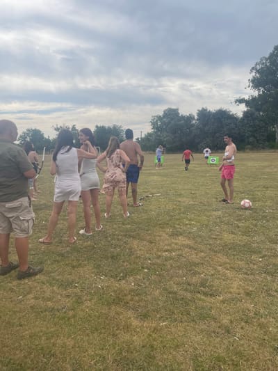 A group of people are gathered on a grassy field under a cloudy sky. Several individuals are standing in a line, while others are dispersed in the background. There is a soccer ball near a shirtless man wearing pink shorts. Archery targets are set up in the distance. The individuals in the foreground appear to be engaged in an archery activity, as one person holds a bow. The overall scene suggests an outdoor social gathering or recreational event.