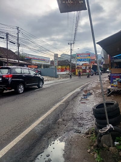 The image captures a vibrant street scene in Cimaung, Indonesia, under an overcast sky, suggesting either an early morning, late afternoon, or a post-rain setting due to the wet asphalt and numerous puddles. The street, showing signs of wear with potholes, features a white dividing line.

In the immediate foreground on the left, a black Toyota Innova SUV is seen driving away from the viewer. The right foreground shows a muddy, unpaved roadside with large puddles reflecting the cloudy sky. A stack of worn tires is tied to a metal pole, next to some rocks and sparse vegetation.

Numerous utility poles crisscrossed with a dense network of power lines line both sides of the road. Further down the street, various commercial buildings and roadside stalls are visible. Prominently featured are several convenience stores: a blue and white Indomaret store on the left, and another Indomaret sign, along with an Alfamart sign, further down on the right. A large billboard with both "Indomaret" and "Alfamart" branding can be seen in the distance, with the faint outline of a mountain behind it.

Other smaller shops and kiosks with colorful fronts are interspersed, including one stall advertising "ES TELER SULTAN 10.000." A tall, white vertical sign with blue text includes "IMVU" and "BRI LINK." Several people are present, including pedestrians walking on the sidewalk or near the road, and motorcyclists navigating the street. An old, partially obscured sign hangs overhead, its text largely illegible. The scene depicts a typical, busy everyday street in an Indonesian town, characterized by local commerce, infrastructure, and daily activity.