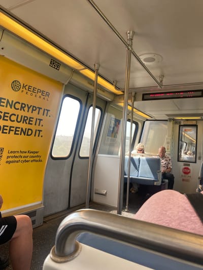 This media file depicts the interior of a train car, likely in Washington, USA. The perspective is from a seated passenger looking forward. Two people are visible: a man with a patterned shirt and a woman with sunglasses, both seated. The train appears to be in motion or stopped, judging by the daylight visible through the windows and the presence of passengers.
Prominent objects include handrails, seats, windows, and advertisements. A large yellow advertisement for "KEEPER FEDERAL" with the slogan "ENCRYPT IT. SECURE IT. DEFEND IT." is featured prominently. Another ad displays "DEALS UP TO 70% TONGER". Signs indicating "STOP", "EXIT", and "DO NOT OPEN" are also visible, along with the car number "3203".
The lighting suggests it is daytime. The overall mood is casual and mundane, typical of public transportation. The scene is a snapshot of daily life on a train.