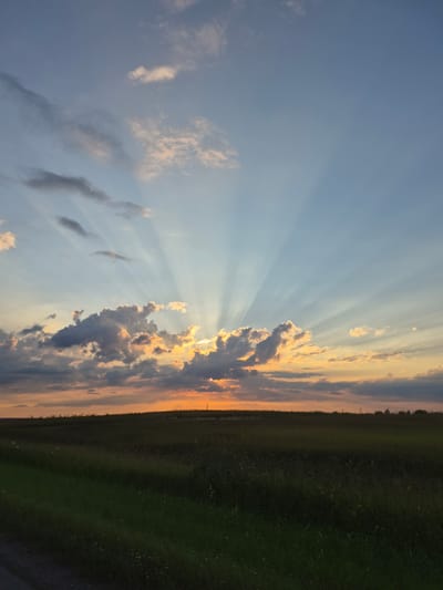 A twilight sky displays crepuscular rays fanning out from behind a bank of dark, dramatic clouds that are illuminated by the setting sun, creating an ethereal glow. The sun itself is mostly obscured by the clouds, with only a hint of its orange light peeking through. Below the sky, a flat, dark expanse of a field stretches to the horizon, with a subtle gradient of green grass in the foreground suggesting a rural or agricultural setting. The scene is devoid of any human presence or structures, creating a sense of vastness and natural beauty. This image was captured in Perevoznoe, Russia.