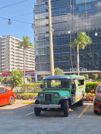 The image shows a green and silver jeepney parked on a concrete surface, likely a parking area. The jeepney has the license plate "DAV 295" and the name "KYRC" on the front windshield. On the jeepney is the inscription "MALIBAY-PASAY ROTONDA-MRT-LRT-SM MOA-LIBERTAD". A red car is partially visible in the foreground. In the background are two modern buildings, one with a glass facade and the sign "NEX GEN TOWER". Palm trees are also visible. The scene is located in Pasay, Philippines. Two workers are visible rappelling down the glass tower.