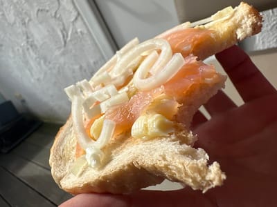 The image shows a close-up of a person holding a slice of bread topped with smoked salmon, thinly sliced white onion, and a dollop of yellow butter or spread. The bread appears to be a rustic loaf with a slightly irregular crust. The salmon is a vibrant orange-pink color and is layered on the bread. The onion slices are translucent and ring-shaped. The background is out of focus but suggests an outdoor setting with a textured wall and wooden decking. The scene is lit by natural light, casting shadows. The photo was taken in Heinebach, Germany.