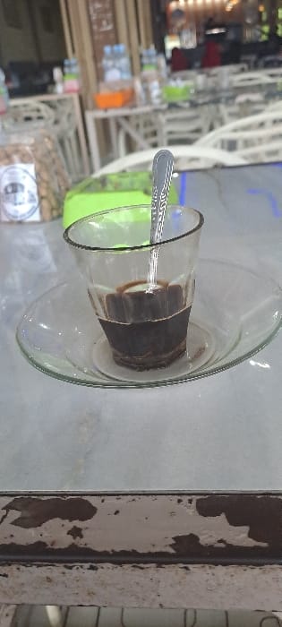 The scene depicts a close-up view within a cafe in Alue Mudem, Indonesia. In the immediate foreground, a clear glass containing dark brown liquid, consistent with coffee, with sediment at the bottom, is centered. A silver-colored spoon is inserted vertically into the liquid. The glass rests on a clear glass saucer, which in turn sits on a light grey or white table surface. Below the table, a horizontal section of a weathered structure with peeling light brown paint revealing darker, possibly metallic, material is visible.
To the left of the glass, a transparent plastic bag containing light brown, irregularly shaped snacks, with a white circular label displaying dark text, is partially visible. Behind the glass, a portion of a bright green plastic container is present. The background is blurred, showing a cafe setting with multiple white, ornate chairs and tables. On one distant table, several plastic bottles of water and an orange-colored beverage are discernible. Wooden architectural elements and indistinct light sources are also present in the background. No people are visible.