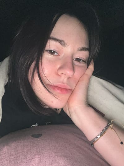 A woman with dark hair is resting her face on her hand while lying on a bed. She has light skin and dark eyes. She is wearing a silver bracelet on her wrist, and her head is tilted to the side. The background includes a pillow and a dark-colored shirt. The lighting is soft, giving the image a relaxed and intimate feel.
