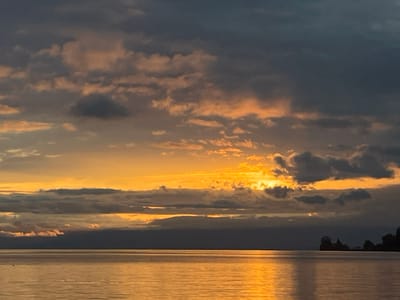 The image presents a panoramic view of Lake Geneva (Lac Léman) in Montreux, Switzerland, at either sunrise or sunset. The sky occupies the upper two-thirds of the frame, characterized by a dynamic cloud formation. Dark grey and blue-grey clouds are prevalent in the upper regions, transitioning to vibrant orange and yellow hues near the horizon, where the sun is partially obscured. A brilliant, golden-yellow light source, presumably the sun, emanates from behind the central cloud cover, casting a strong illumination. The lower third of the image is dominated by the calm waters of Lake Geneva, which reflect the sky's warm tones, appearing golden-orange with subtle ripples visible across its surface. On the far right, a dark, indistinct landmass or series of structures is silhouetted against the bright horizon. There are no discernible individuals, specific objects, or actions within the scene.