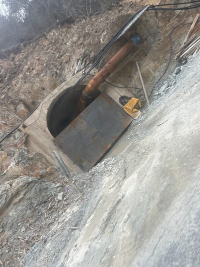 This image captures a construction or infrastructure installation site in Erma Reka, Bulgaria, centered on a tunnel entrance into a steep, rocky hillside. The tunnel, appearing circular and concrete-lined, features a large, rusty, brown metal pipe emerging from its interior and extending downwards at an angle towards the right. This pipe has a blue cap or fitting on its visible end outside the tunnel. A dark grey, rectangular metal structure, possibly a cover or access panel, is positioned over the lower section of the tunnel opening. Multiple black electrical cables run along the hillside and near the tunnel entrance, with some routed over a corrugated metal sheet acting as a partial overhead cover. To the right of the metal structure, a crumpled yellow tarp or fabric is visible on the ground, alongside a scattered pile of wooden planks and debris. The ground in the foreground and lower right consists of gravel and dirt, forming a sloped path or work area, with evidence of moisture and uneven terrain. The upper background reveals a rocky, treeless slope with bare deciduous branches under an overcast sky. No individuals are present in the scene.