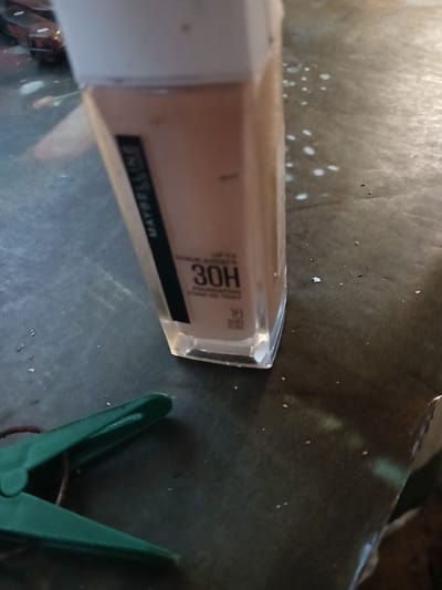 The image shows a close-up of a bottle of Maybelline 30H foundation on a dark, textured surface. The bottle is mostly in focus, with some scattered white speckles on the surface. A green clothespin is visible in the bottom left corner of the frame. The scene appears to be indoors.