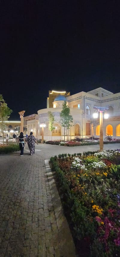 The image depicts a night scene in what appears to be an outdoor plaza or courtyard in the city of Satkak, Uzbekistan. In the foreground, a cobblestone pathway leads into the scene, bordered by a vibrant flowerbed filled with a variety of colorful blooms, including white, yellow, pink, and purple flowers. Two women are walking away from the camera on the pathway, one wearing a dark top and pants, and the other in a patterned, flowing dress.
Behind them, a building with distinctive architectural features is illuminated. The building is constructed with a light-colored stone or stucco, adorned with intricate blue and white patterned tiles. A prominent blue dome sits atop a section of the building, and arched windows with latticed screens are visible. To the left of the dome, a modern, curved building with illuminated windows rises higher. Several lampposts with multiple lights are strategically placed, casting a warm glow on the pathway and surrounding area. In the background, additional illuminated architectural elements and trees can be seen, contributing to the overall ambiance of the scene. The sky is dark, indicating it is nighttime.