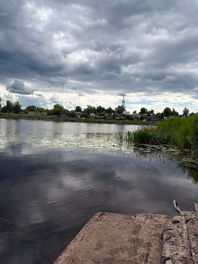 The image depicts a serene river scene in Kimry, Russia, under a dramatic, cloud-filled sky. In the foreground, a concrete structure, possibly a dock or landing, occupies the lower right corner. The surface of the dark water reflects the cloudy sky and the distant shoreline. The river is dotted with lily pads and some of their yellow flowers. Along the riverbank, a cluster of traditional wooden houses and a white church with a golden dome are visible, nestled amongst lush green trees. Tall reeds and grasses grow along the right bank of the river, adding to the natural beauty of the landscape. The overall atmosphere is tranquil, with a sense of rural Russian charm.