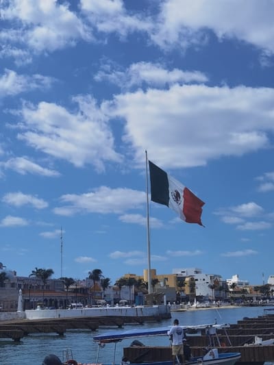 The image shows the Mexican flag flying on a tall flagpole in the city of Cozumel, Mexico. The flagpole is located in the middle of the image and stands in front of a waterfront promenade. There is a man standing on the promenade to the right of the flagpole wearing a blue t-shirt, black shorts, and a white baseball cap. To the left of the flagpole, there are many small boats docked along the waterfront. The background of the image shows a clear blue sky with white clouds. The scene is shot from a distance, looking towards the flagpole. The location is approximately 20.512° N, 86.95° W, which is near the downtown area of Cozumel.