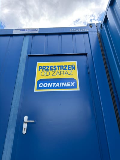 The image shows the side of a blue shipping container. A white sign with yellow lettering is affixed to the container door. The sign reads "PRZESTRZEN OD ZARAZ" at the top and "CONTAINEX" at the bottom. The container has a silver door handle and a lock. In the background, there is a cloudy sky. The container is located in Stróża, Poland.