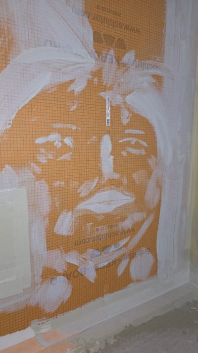 The image depicts a close-up, vertical view of a shower wall under construction in Bremerton, Washington, United States of America. The wall is covered with a textured orange membrane, possibly a waterproofing material, imprinted with a grid pattern and manufacturer's text including "www.schluter.com". White adhesive or waterproofing compound has been applied in broad strokes, obscuring portions of the membrane and creating an abstract, face-like pattern. The face appears to be formed by white paint or caulk on the orange surface, with visible features such as eyes, nose, and mouth suggested by the application of the material. The grid lines on the membrane serve as a reference for scale and alignment. The bottom of the image shows the edge of the shower curb and the floor, both of which are coated with a light gray material, likely a waterproofing membrane or leveling compound. The overall scene suggests an ongoing bathroom renovation or construction project.