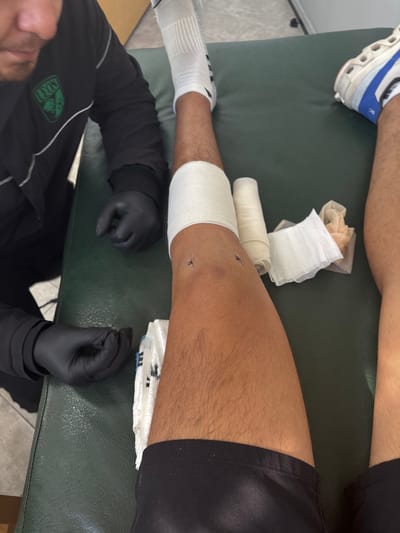 A patient lies on a green examination table. Their lower leg and knee are wrapped in white bandages, with stitches visible. Medical supplies, including gauze and tape, are scattered nearby. A medical professional, wearing a black long-sleeve shirt and black gloves, attends to the patient, their face partially visible in the lower left corner. The patient wears a white sock and a white and blue sneaker. Black shorts are visible around the upper thigh area.