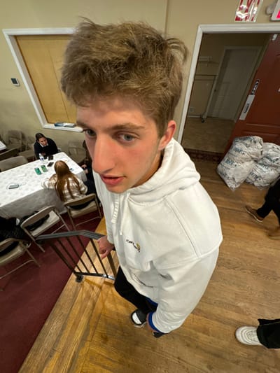 An indoor scene in Bergenfield, United States, depicts a young male in the immediate foreground, looking directly at the camera from a downward angle. He has light brown, tousled hair, blue eyes, and is wearing a white Carhartt hoodie, dark pants, and black and white slides with blue socks. His left hand is on a black metal stair railing.
In the mid-ground to the left, several individuals, predominantly young females, are seated around a white-tablecloth-covered table with folding chairs on a red carpeted floor. Visible on the table are a plastic water bottle, green cans, and cell phones. The room features light-colored walls with a window-like opening covered by tan blinds.
To the right, a partially open reddish-brown door reveals an interior room with light-colored walls and a tiled floor. Adjacent to the door, on the wooden floor, are multiple clear plastic bags filled with white, rolled materials, labeled "TOPEK 100% MICROFIBER." The lower legs and feet of at least two other individuals are visible near these bags; one wears brown shoes, another white sneakers. An illuminated "EXIT" sign is affixed above the doorframe. The overall lighting is artificial and bright.
