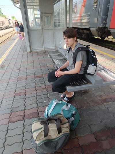 A young person with dark hair is sitting on a metal bench at a train station, looking towards the camera with a neutral expression. They are wearing a grey t-shirt, black pants, and black slide sandals with white logos. A black backpack with grey accents is on their back. Two duffel bags, one teal and one beige with dark green trim, are placed on the patterned concrete ground in front of the bench. In the background, two women are walking away from the camera on the train platform, one pulling a pink suitcase. A train with red and grey accents is parked beside the platform. The environment appears to be an outdoor train station with a covered waiting area.