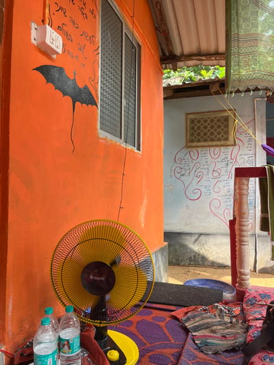 The image captures an outdoor or semi-outdoor area in Gokarna, India, between two structures. The foreground features an orange stucco wall on the left, displaying a white electrical switchboard with two switches and one socket, below which are black painted Hebrew script and a black bat silhouette with an elongated tail. An aluminum-framed window with textured privacy glass is set into this wall. On the ground directly below, a black and yellow "ORIENTAL" brand pedestal fan rests on a purple and orange patterned mat, flanked by two clear plastic Bisleri water bottles and a folded red fabric.

To the right, an open space reveals a light blue wall, adorned with red painted swirls, various scripts (some resembling Hebrew), and a small sun motif. A framed rectangular object, possibly a ventilation grid or decorative panel, hangs on this blue wall. A reddish-pink wooden pillar with white peeling paint stands in front of this wall. Overhead, a corrugated metal roof is visible, with green foliage beyond it. A green patterned textile or curtain and a yellow cord are suspended across the upper right portion of the scene. The ground is a sandy or dusty light brown surface. No individuals are present.