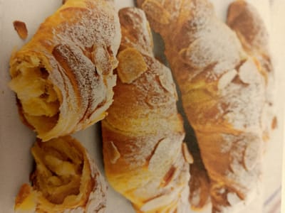 The image displays a close-up, overhead view of several golden-brown croissants, generously dusted with powdered sugar and adorned with slivered almonds. The croissants are arranged in a cluster, with some overlapping. The texture of the flaky pastry is evident, and the lighting suggests a warm, possibly indoor setting. There are no people or distinct environmental features visible in the frame. The image is a still life focusing solely on the baked goods. The provided coordinates, Rumaithiya, Kuwait, do not correspond to any discernible elements within the visual content of the image itself.