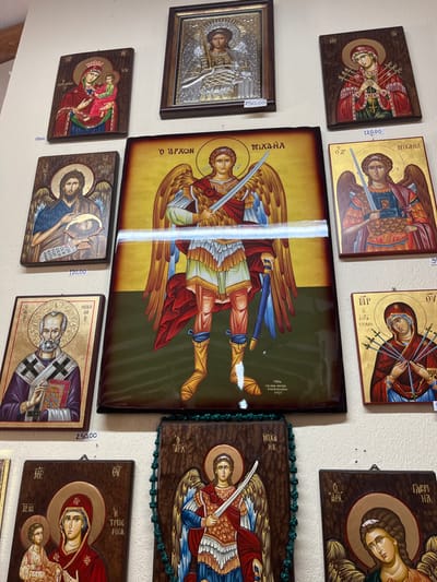A wall in a shop in Moni Archaggelou, Greece, located at the coordinates 40.597, 24.702, is decorated with nine framed religious icon paintings. The icons depict various religious figures such as angels, saints, and the Virgin Mary. The paintings are framed in a variety of woods and styles. They are hung at different heights and angles, some with price tags attached.  The wall is painted a light yellow color. In the center of the image is a framed icon of the Archangel Michael, a popular subject in Greek Orthodox art. This icon is surrounded by other icons of angels, saints, and the Virgin Mary.  A string of green beads hangs from the upper right corner of the image, likely from the adjacent shop counter. The icons appear to be for sale in the shop.