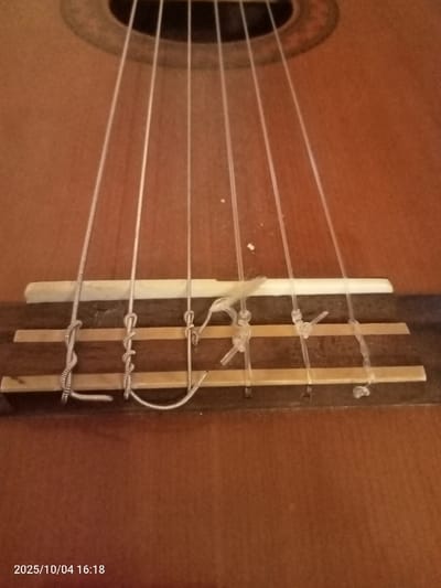 This is a close-up, static photograph of the bridge and lower soundboard area of a classical or acoustic guitar, taken in Htongyidaung, Myanmar (Burma). The instrument features a light-brown wooden soundboard and a dark wooden bridge. A white, rectangular saddle is positioned on the bridge. Six strings are visible: three silver-colored wound bass strings on the left, and three translucent white/clear treble strings on the right, all extending upwards towards the partially visible sound hole with its circular rosette at the top. The strings are secured to the bridge. The three bass strings are tied with coiled knots around the bridge tie-block; specifically, the third bass string from the left displays a loose, U-shaped end protruding beneath its knot. The three treble strings are also tied to the bridge, exhibiting less uniform knotting, with the second treble string from the right showing a frayed end emanating from its securing knot. Minor light-colored debris or dust particles are present on the soundboard above the bridge. No people, active subjects, or explicit actions are depicted. The timestamp 2025/10/04 16:18 is visible in the lower-left corner.