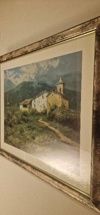 A photograph captures a framed picture mounted on a plain, light-colored wall, observed from an upward, oblique angle. The frame is ornate, featuring a textured, metallic finish with mottled gold and silver tones, encompassing an inner golden fillet and a wide, off-white mat. The artwork, viewed through glass reflecting ambient light, depicts a rural mountainous landscape.
In the midground of the artwork, a winding dirt path traverses a field of varied green and brown vegetation, leading towards a cluster of old buildings. The main structure on the right is stone-built with a prominent square bell tower and a pitched roof, consistent with a small church or chapel. An adjacent, smaller, whitewashed building with a darker roof is positioned to its left. A rustic wooden fence is partially visible near the right side of the path. The background presents steep, dark green mountains partially obscured by heavy grey-blue clouds or mist. No human figures or discernible actions are present within the artwork or the observable environment. This scene is physically located in the city of Blaye, France.
